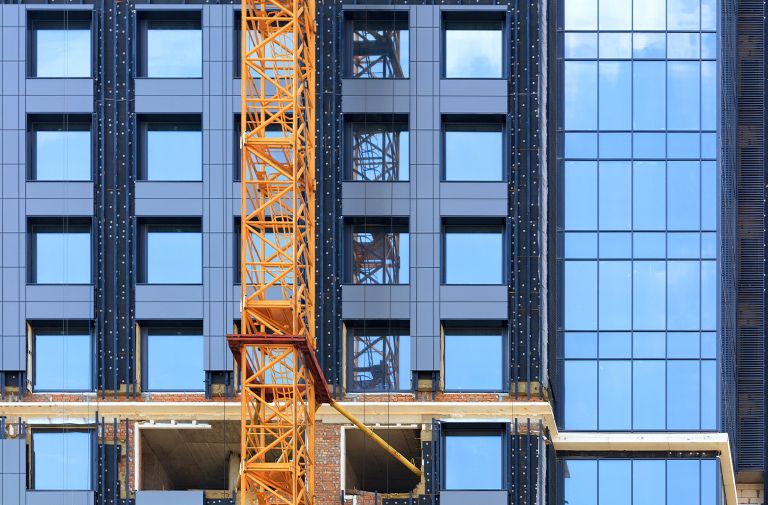 A fragment of the facade of a house under construction with a crane and blue sky reflected in glass